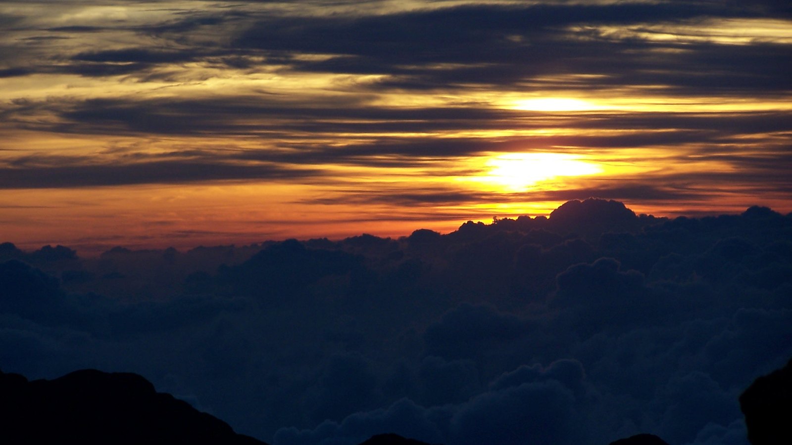 Sunrise light above a sea of clouds and the dark summit landform of Haleakalā