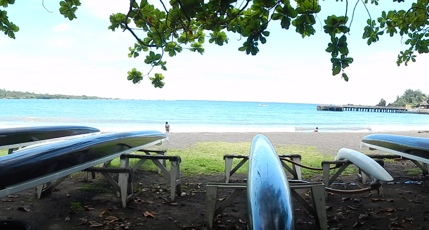 Outrigger canoes stored along the shore of Hāna Bay with the water and pier beyond