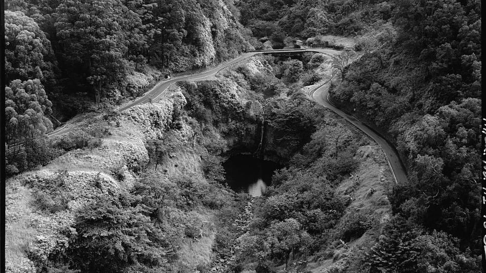 Historic aerial view of Hāna Belt Road curving around Kōpiliʻula Bridge, a waterfall, and a pool