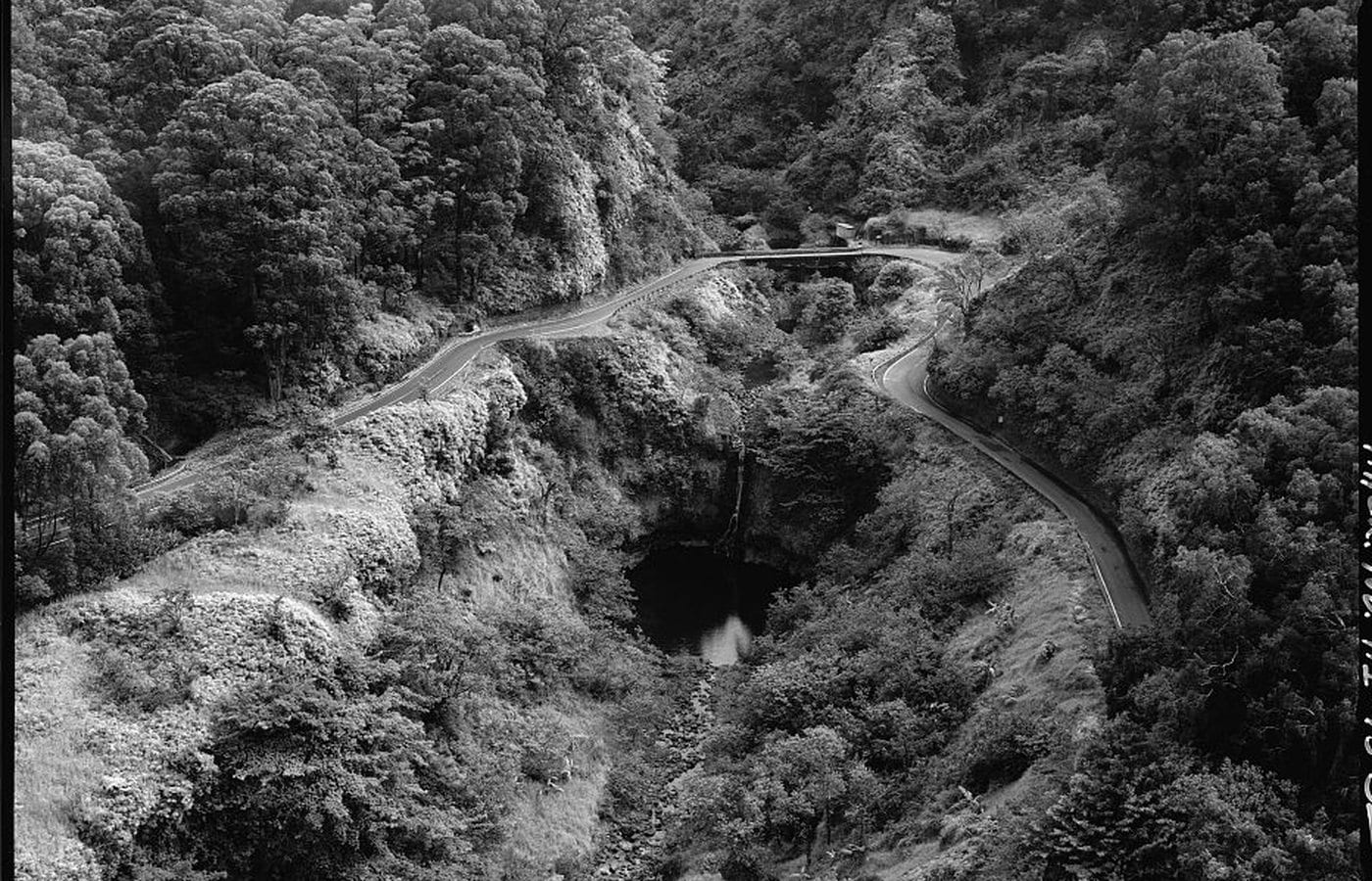 Historic aerial view of Hāna Belt Road curving around Kopiliʻula Bridge, a waterfall, and a pool